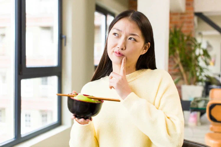 An image of a woman with a ramen noodle bowl in one hand and a confused face. 